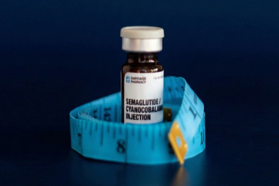 Brown glass vial with white cap labeled 'Semaglutide/Cyanocobalamin Injection' nestled in a blue measuring tape on a dark blue background.