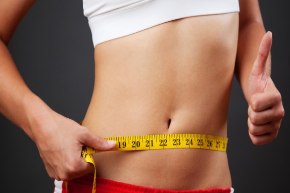 Person in sportswear measures their waist with a yellow measuring tape and gives a thumbs-up gesture around the midsection.