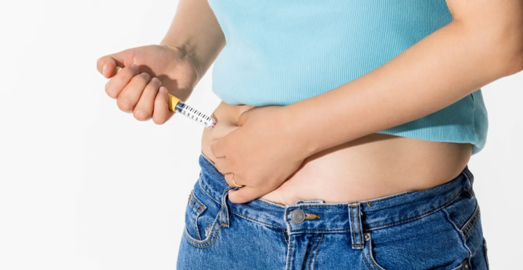 Person injecting a syringe into their abdomen (likely insulin self-injection) while wearing a blue shirt and jeans, indicating diabetes management.