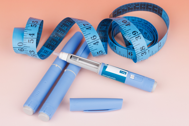 Blue insulin pen with its cap removed, two blue lancets or pens beside measuring tapes, and a blue measuring tape coiled on a pink background, representing diabetes management tools