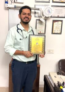 Male doctor with a stethoscope shows a gold-framed award in his clinic, with certificates on the wall behind him.