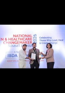 Three people pose on stage during an awards ceremony, with the center man holding a framed certificate and a trophy against a banner that reads National Health & Healthcare ChangeMaker Summit & Awards 2026.