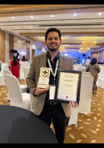 Man in a beige suit posing with a framed certificate and a trophy at an awards ceremony in a decorated conference hall, wearing a lanyard badge.