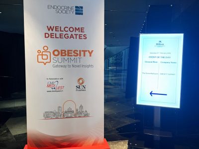 Tall white conference banner reading 'Welcome Delegates' for the Obesity Summit with Endocrine Society logo and a skyline graphic, beside an illuminated Hilton directory sign.