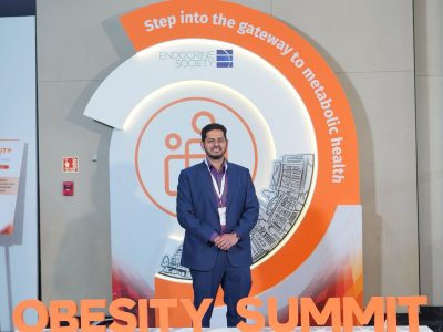 Man in a blue suit stands in front of an orange circular summit display reading OBESITY SUMMIT, with 'Gateway to Novel Insights' banner below.